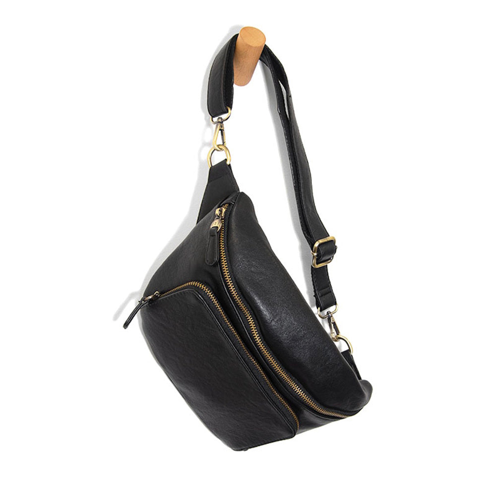 Black leather crossbody fanny pack with gold zippers hanging on a wooden wall peg, stylish waist bag for travel.