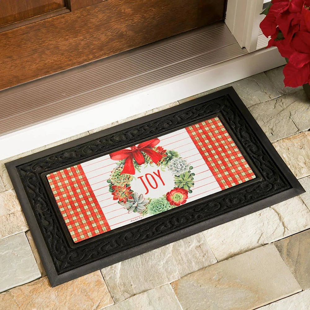 Decorative doormat with a succulent wreath, red bow, and "JOY" text, framed by a black scrollwork border and plaid accents.