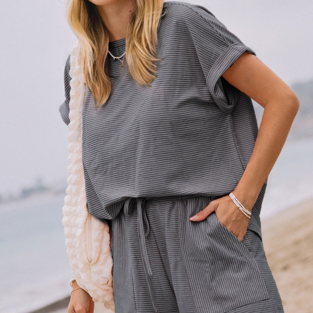 A woman on a beach wearing a grey, finely striped short-sleeve top and matching drawstring shorts set.