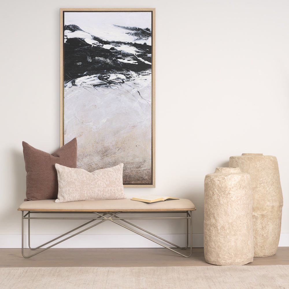 Lifestyle view of the framed abstract canvas art hung above an upholstered bench styled with pillows, a book, and large textured floor vases.