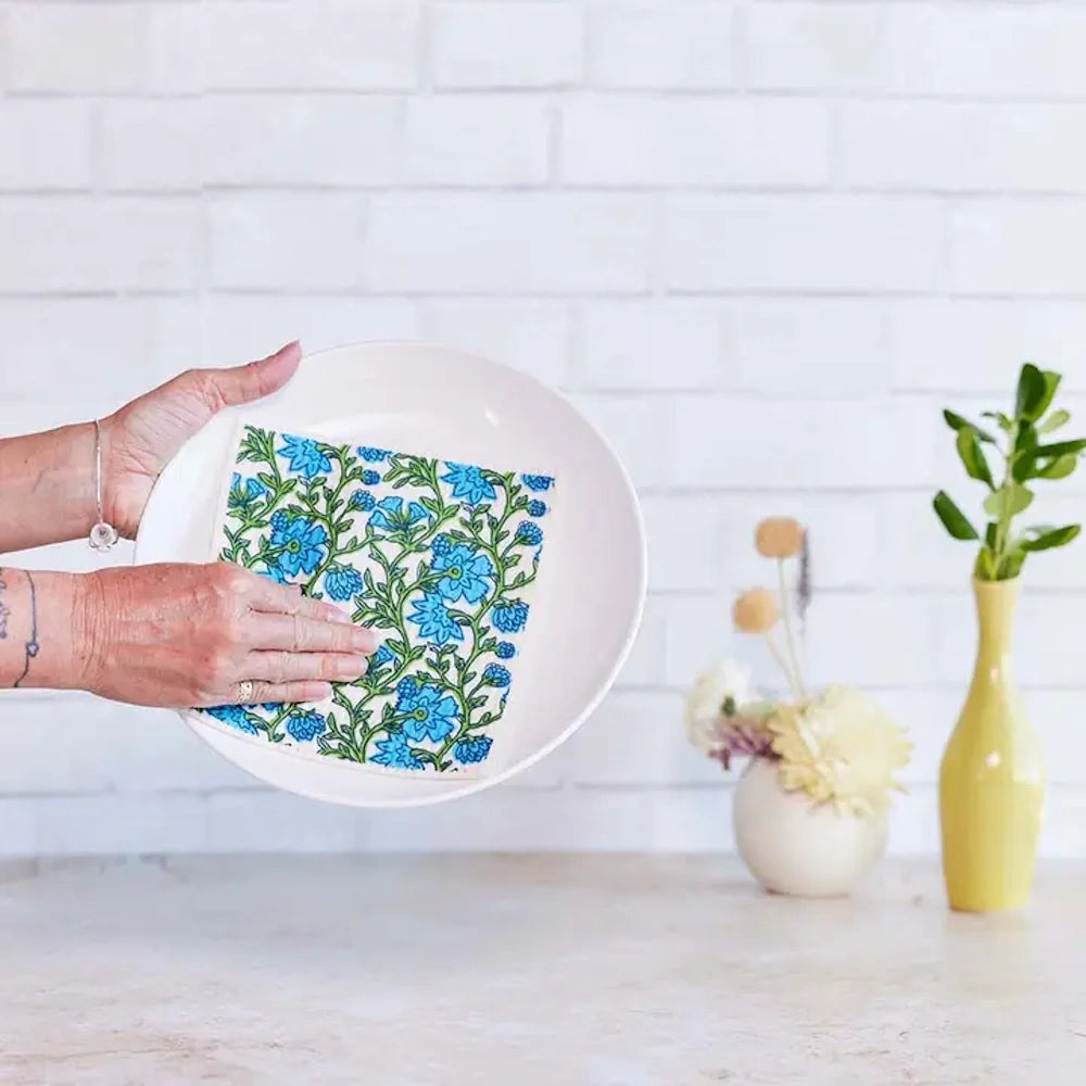 Person drying a white ceramic plate with a floral-patterned blue and green Blu Eco sponge cloth against a white brick wall.