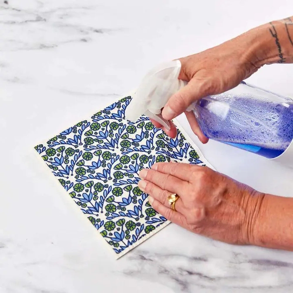 Hands spraying cleaning liquid onto a patterned blue and green eco-friendly sponge cloth on a marble surface.