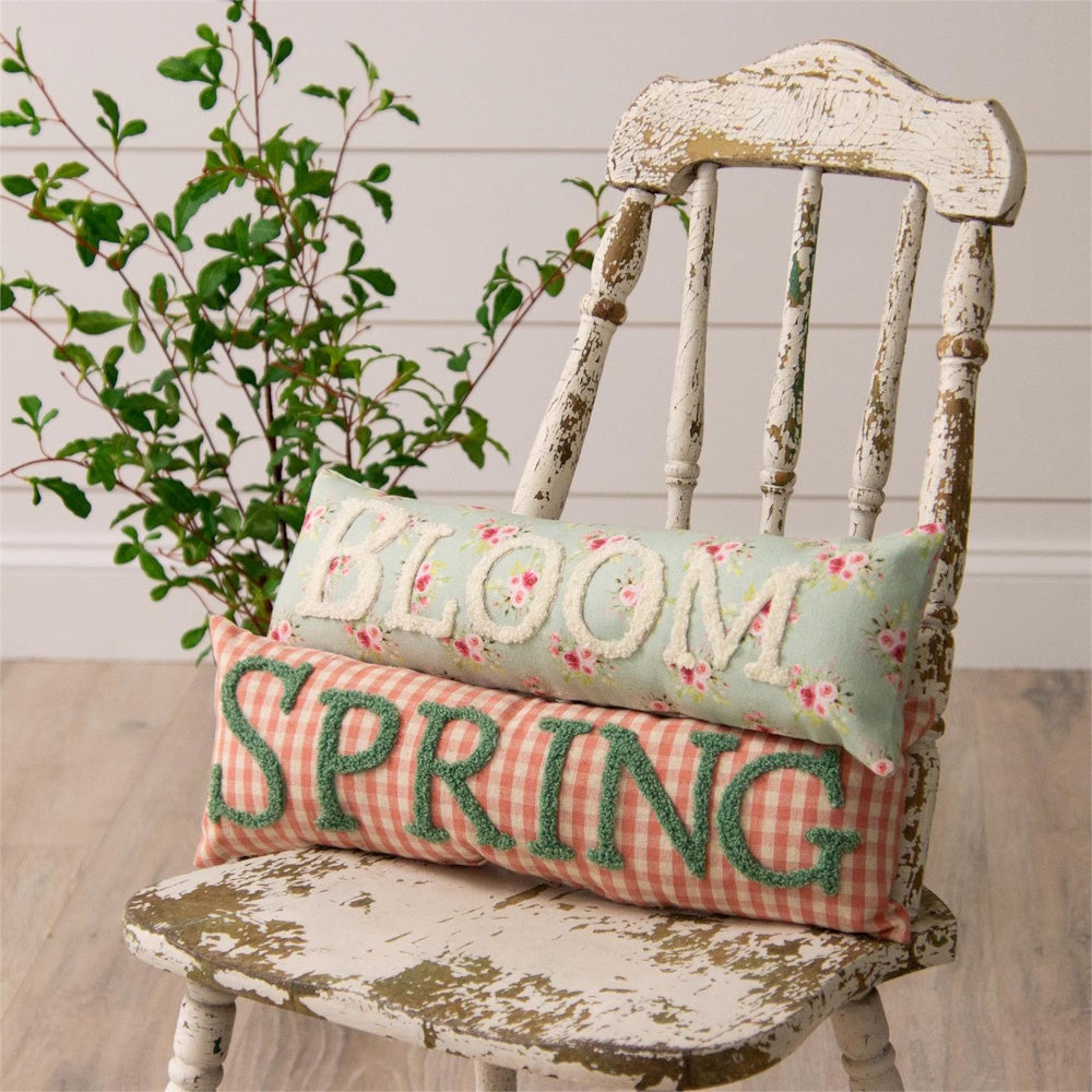 Two decorative spring pillows reading BLOOM and SPRING stacked on a rustic, white wooden chair.