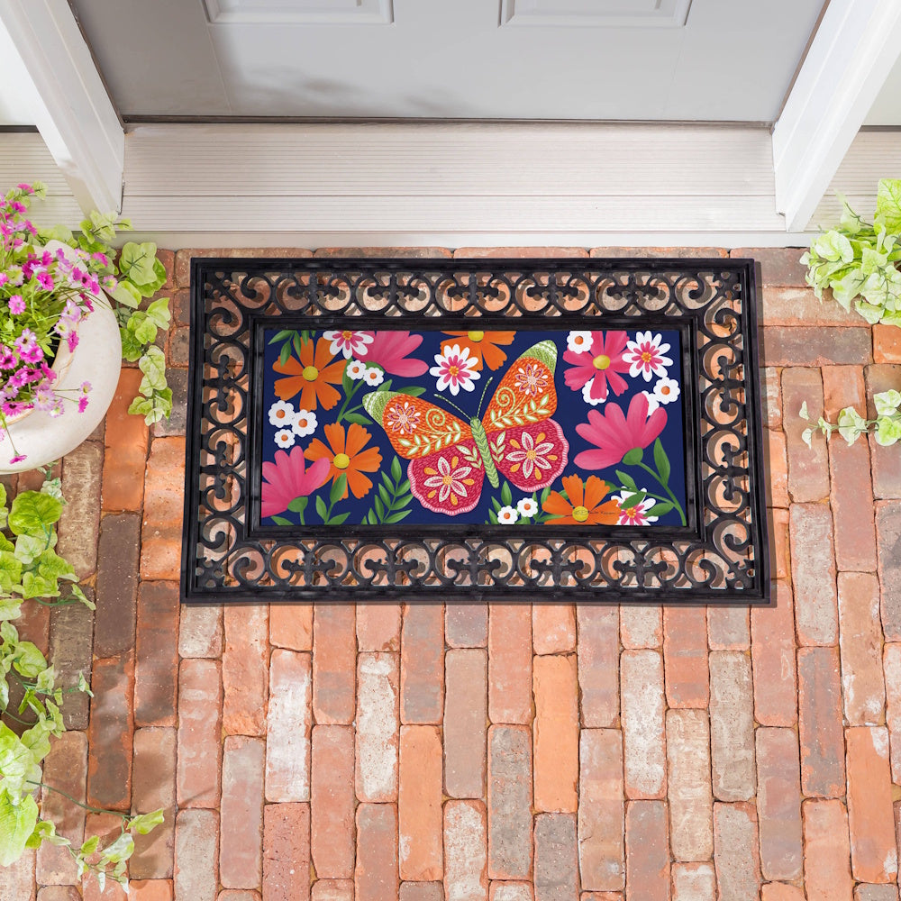 Colorful butterfly and floral doormat insert in a black scrollwork frame on a brick porch, decorative garden home decor.