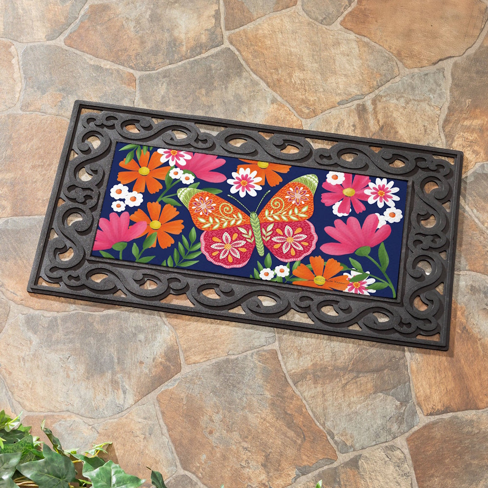 Bloom Burst butterfly and floral Sassafras switch mat in a black scrollwork frame on a stone patio.