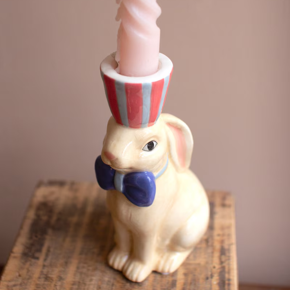 Ceramic bunny taper candle holder with a blue bowtie and striped hat, holding a pink twisted candle on a wooden surface.