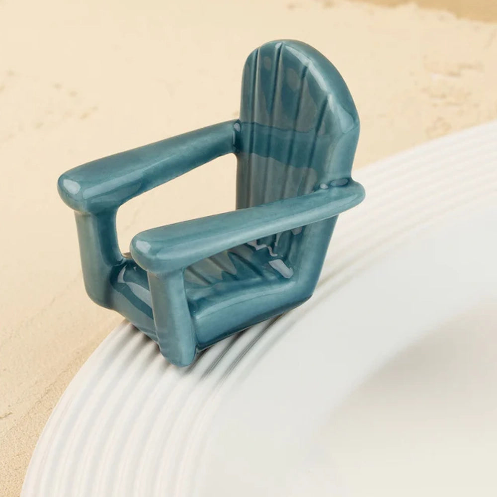 A miniature, glossy blue ceramic Adirondack chair ornament sitting on the rim of a white plate.