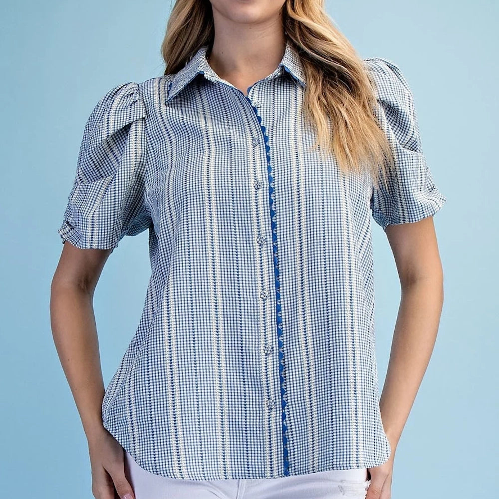 Woman in blue embroidered gingham button-down shirt with ric-rac trim, puff sleeves, and a pointed collar against blue.