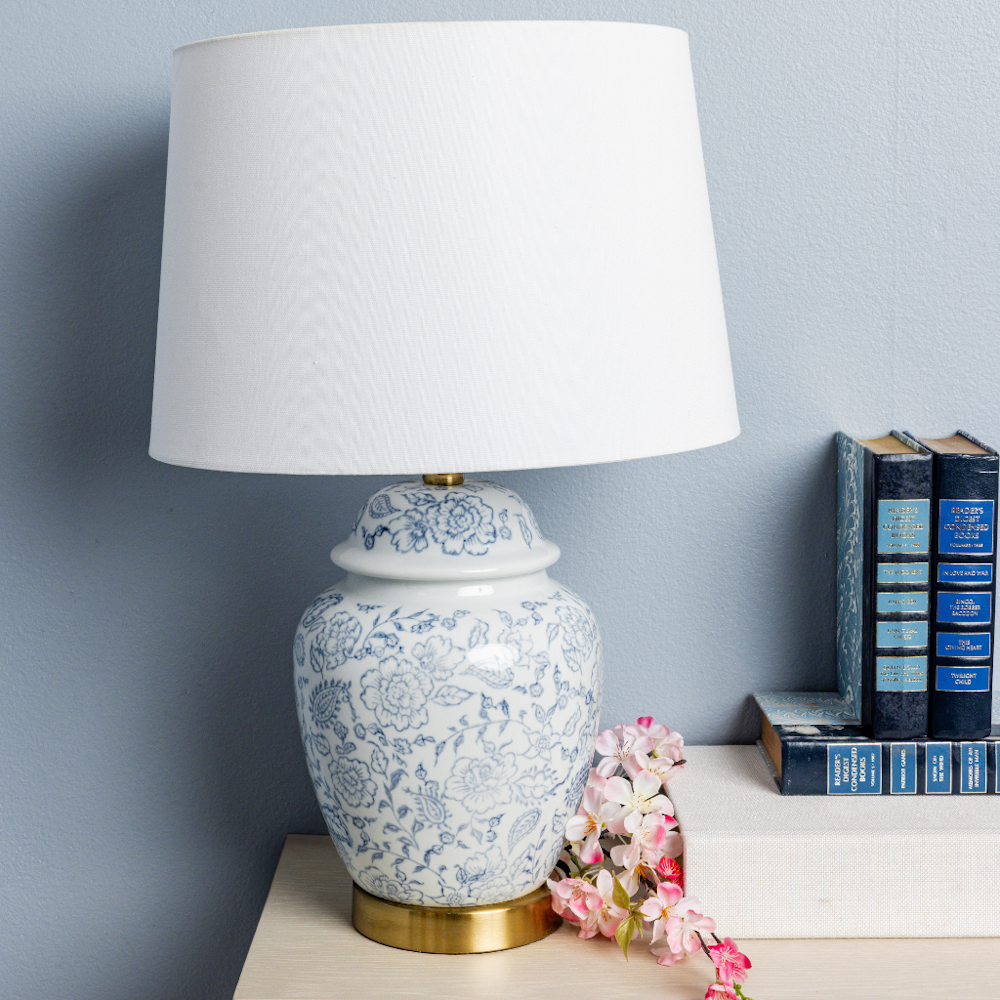 Blue and white floral ginger jar table lamp with white drum shade and gold base on a nightstand with books and pink flowers.