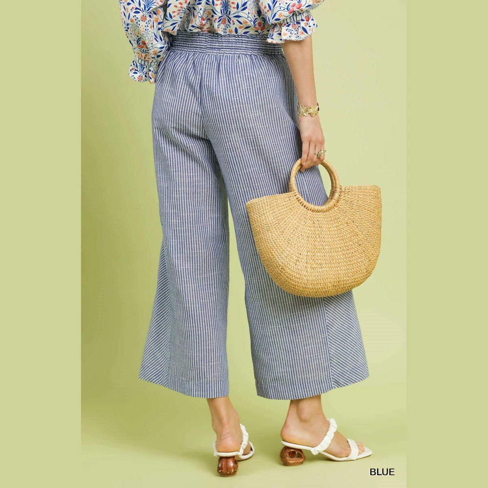 Blue striped wide-leg linen pants with an elastic waist, paired with white sandals and a straw bag.