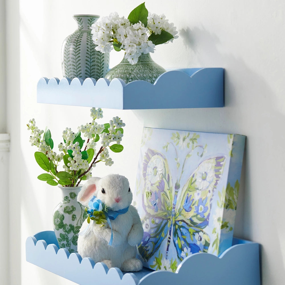 Two light blue scalloped floating shelves holding white flowers in green vases, a bunny figurine, and butterfly art.
