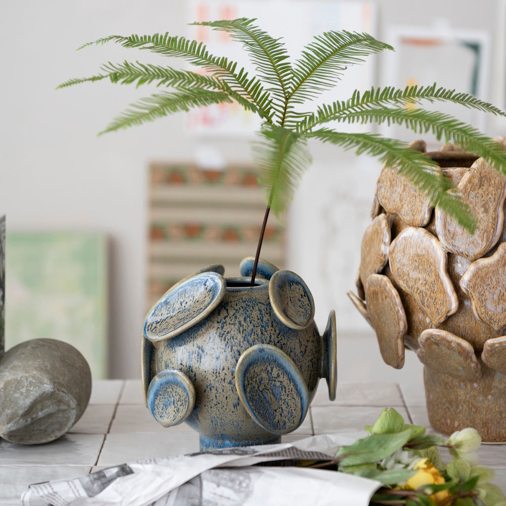 Blue stoneware sphere vase with disc details, holding a single fern frond next to a textured brown vase.