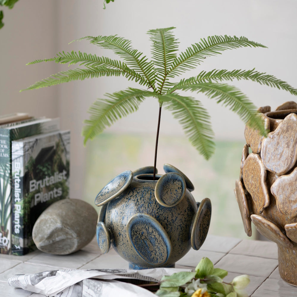Blue speckled round ceramic vase with disc accents holding a single green fern on a tiled surface.