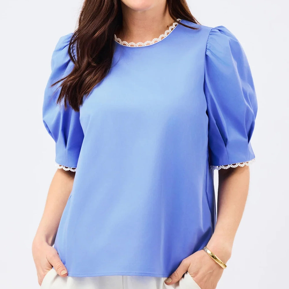 A woman wearing a blue short-sleeve top with puff sleeves, white lace trim, and her hands in her pockets.