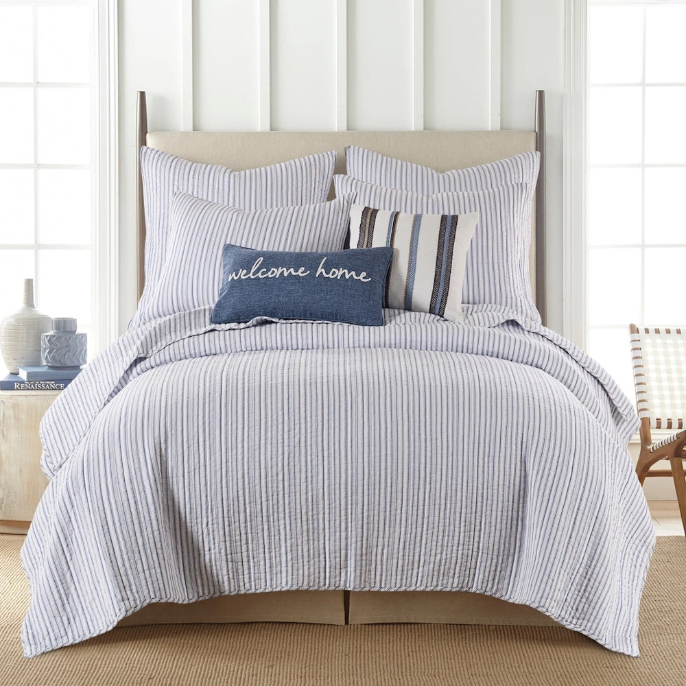 Blue and white striped quilt set on a bed with a "welcome home" accent pillow in a bright, coastal-style bedroom.