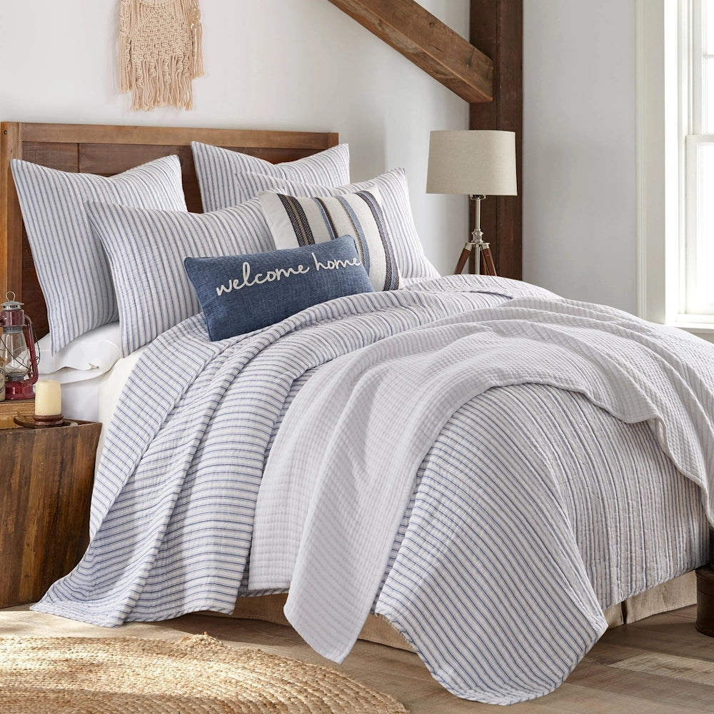 Blue and white striped quilt set on a wooden bed with a "welcome home" accent pillow in a rustic bedroom.