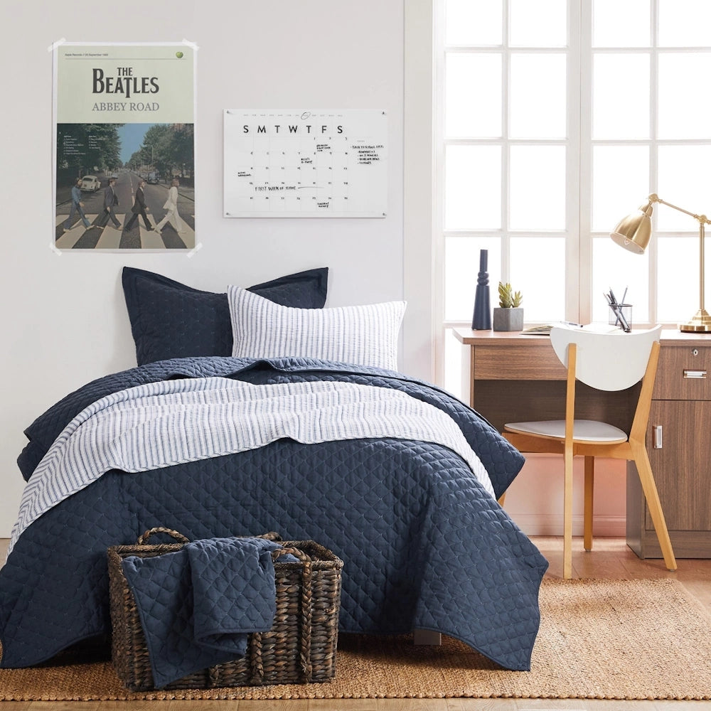 Navy blue quilted bedding set with striped accent pillows in a modern bedroom with a wooden desk and Abbey Road poster.