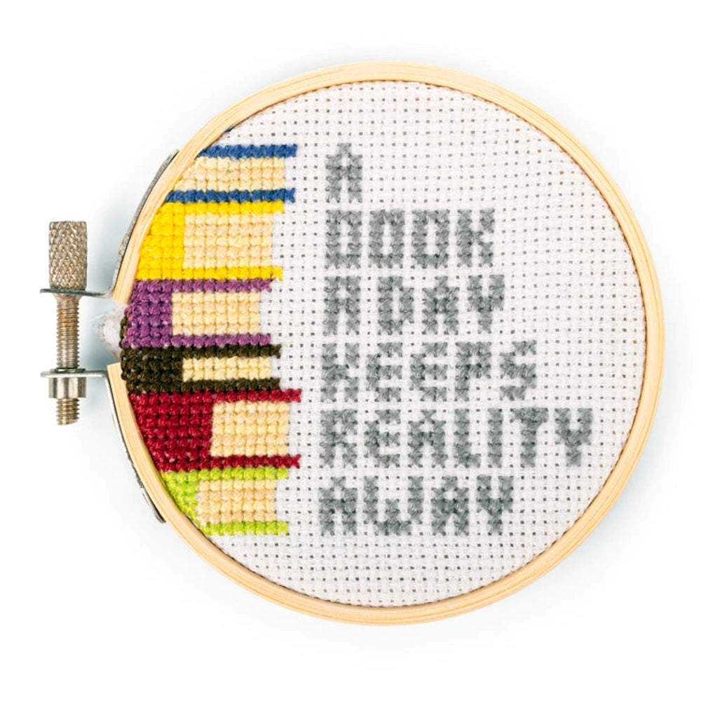 Cross-stitch in a wooden hoop showing a stack of colorful books and the text, "A book a day keeps reality away."