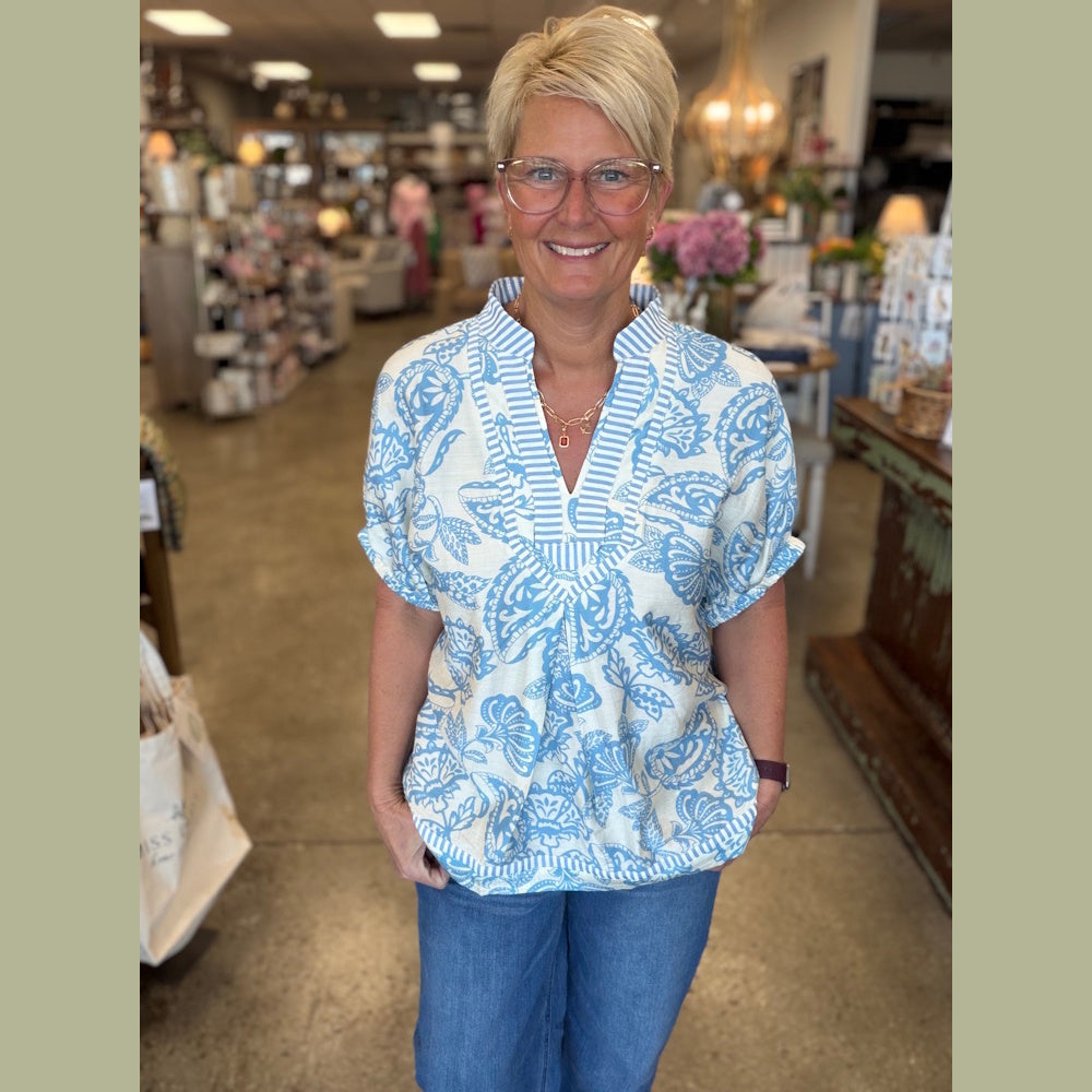 Woman with short blonde hair smiling in a boutique, wearing a blue and white patterned tunic top and jeans.