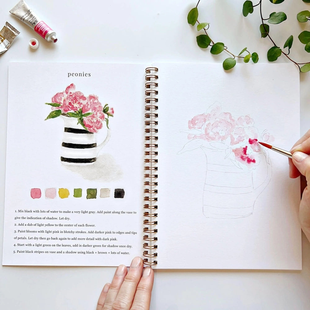 A hand painting pink peonies in a striped vase in an open sketchbook with watercolor paint tubes nearby.