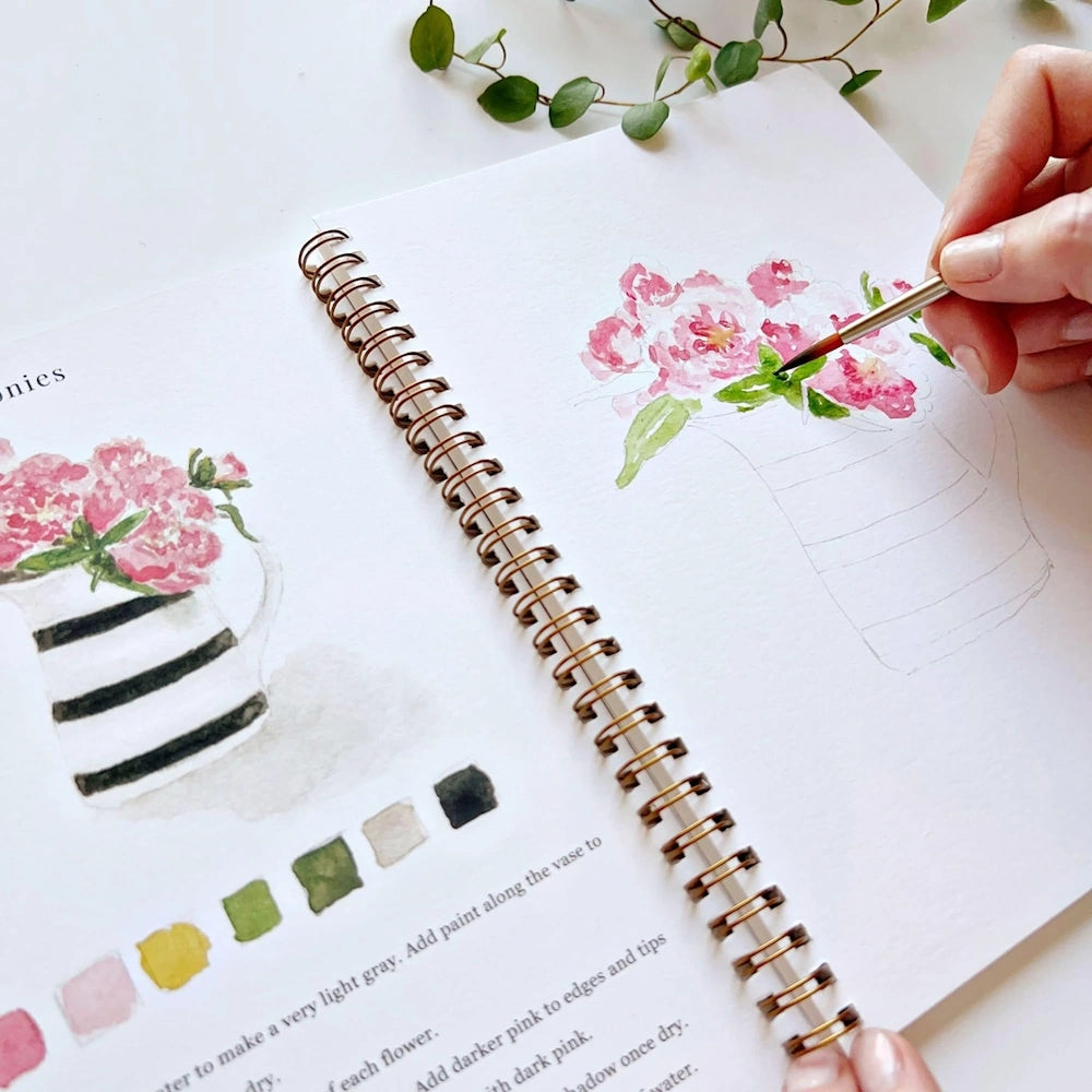 Hand painting pink flowers with a paintbrush in a spiral sketchbook.