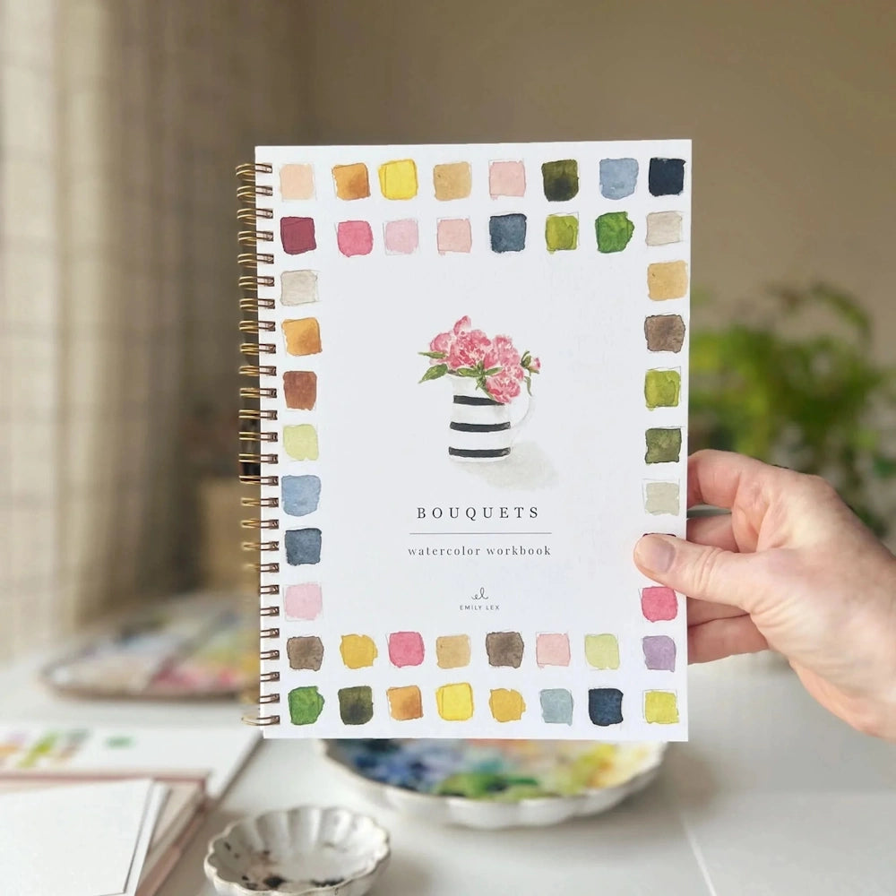 A hand holding a "Bouquets" watercolor workbook by Emily Lex with colorful swatches and a flower illustration.
