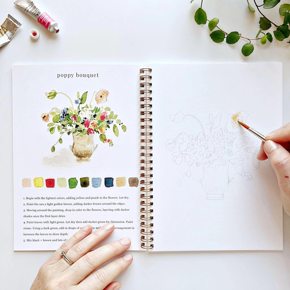 A person paints a watercolor poppy bouquet in a spiral notebook next to a finished illustration and guide.