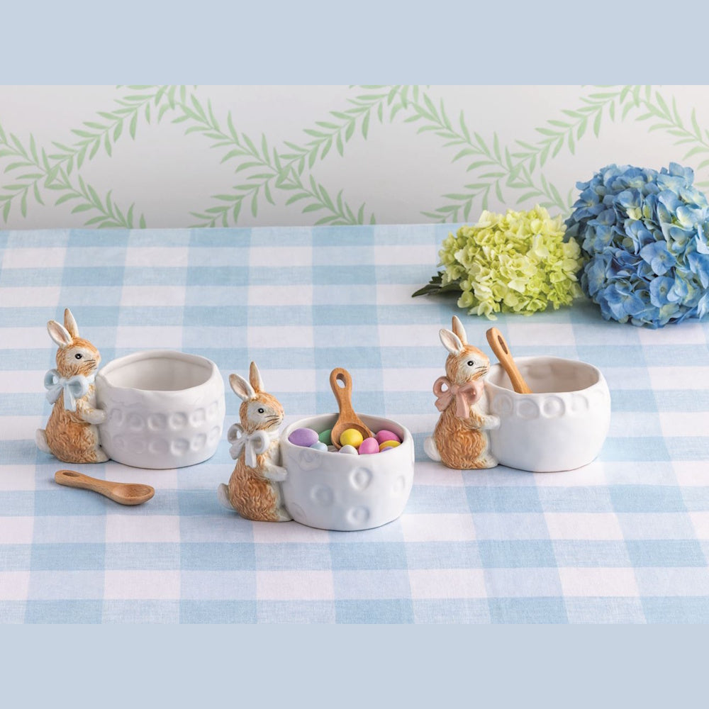 Three ceramic bunny candy dishes with bows and wooden spoons on a blue checkered tablecloth with Easter candy.