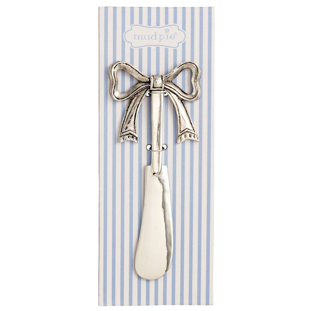 Silver metal cheese spreader with a decorative bow handle, displayed on blue and white striped Mud Pie packaging.