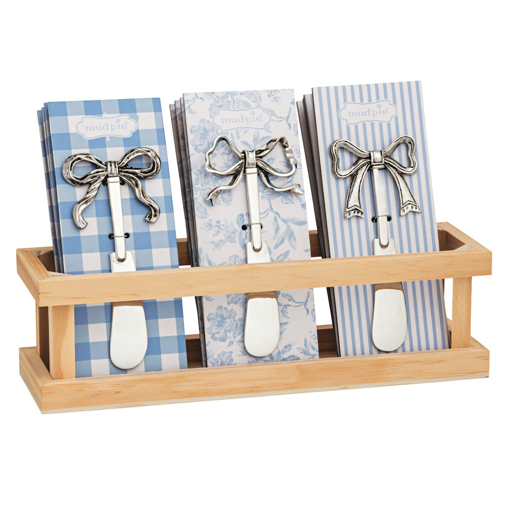 Three silver bow-topped cheese spreaders displayed on blue patterned cards in a wooden crate holder.