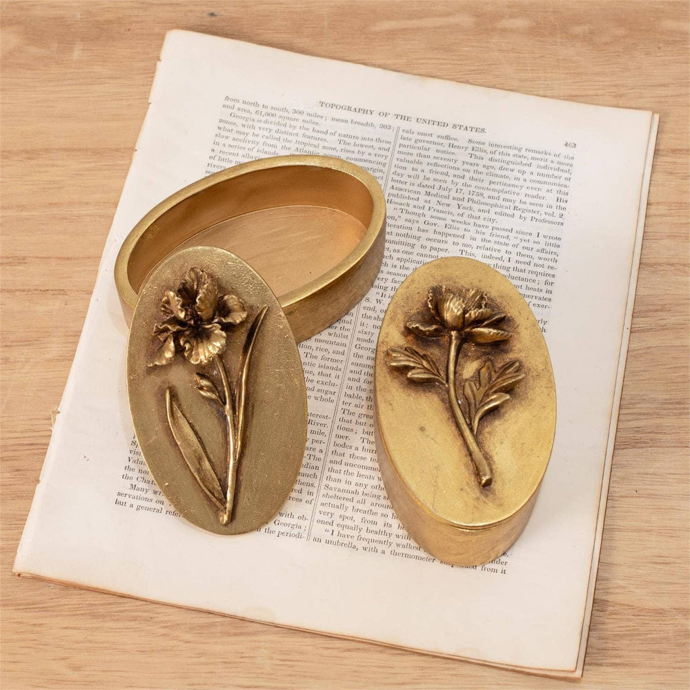Pair of vintage brass floral trinket boxes with embossed iris and poppy lids on an antique book page.