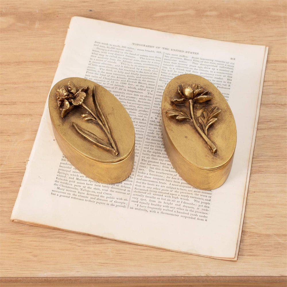 Pair of vintage brass-tone oval trinket boxes with raised floral iris and tulip designs on an antique book page.