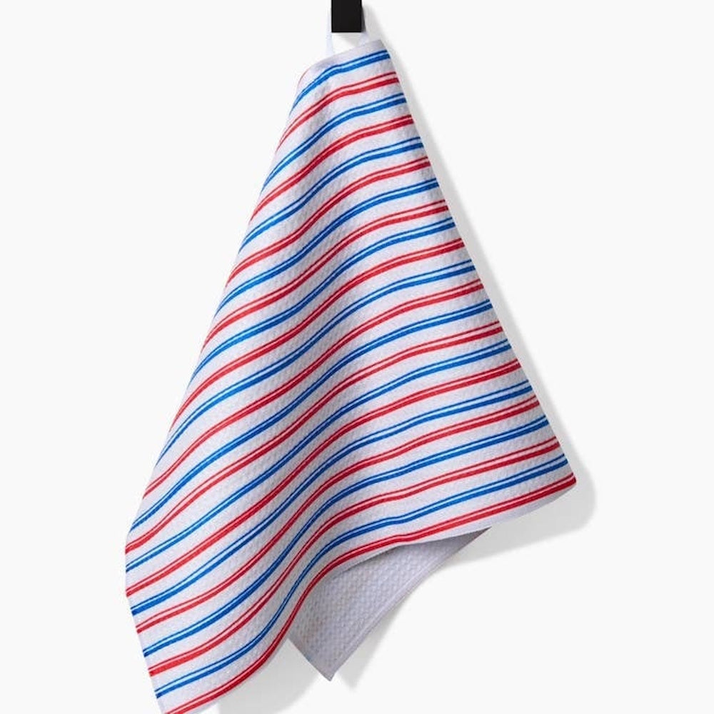A white bar towel with thin, repeating red and blue diagonal stripes, hanging from a black hook against a white wall.