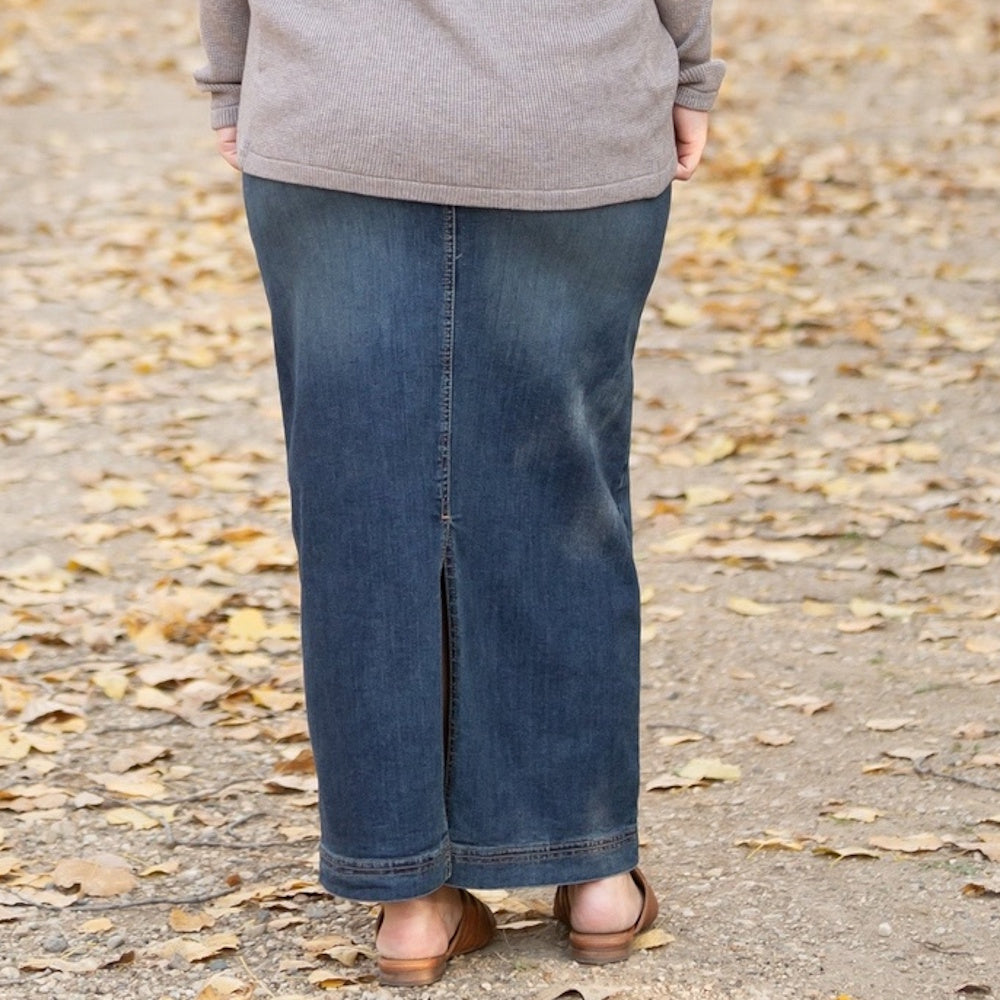 Back view of a person standing outdoors wearing a long, dark wash denim maxi skirt with a back slit.