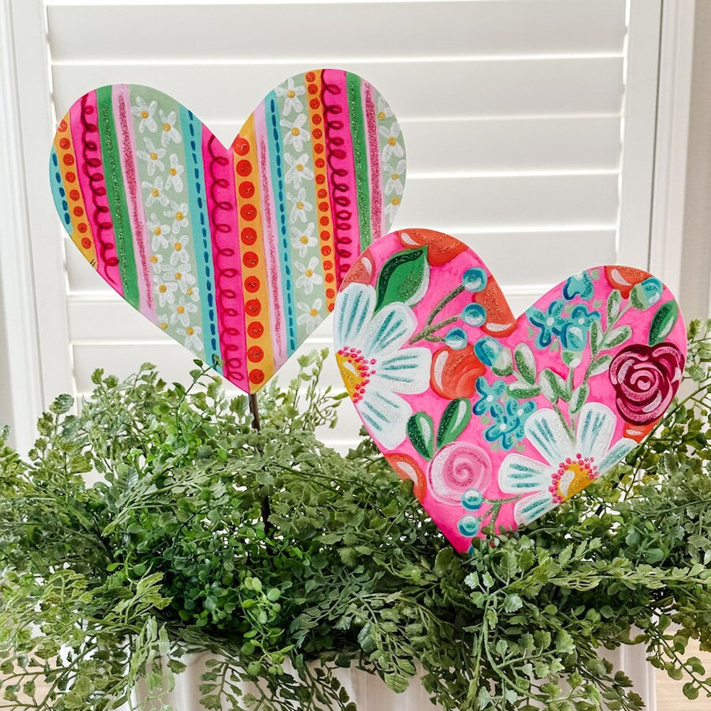 Two vibrant hand-painted heart garden stakes with floral and striped patterns tucked into green ferns.