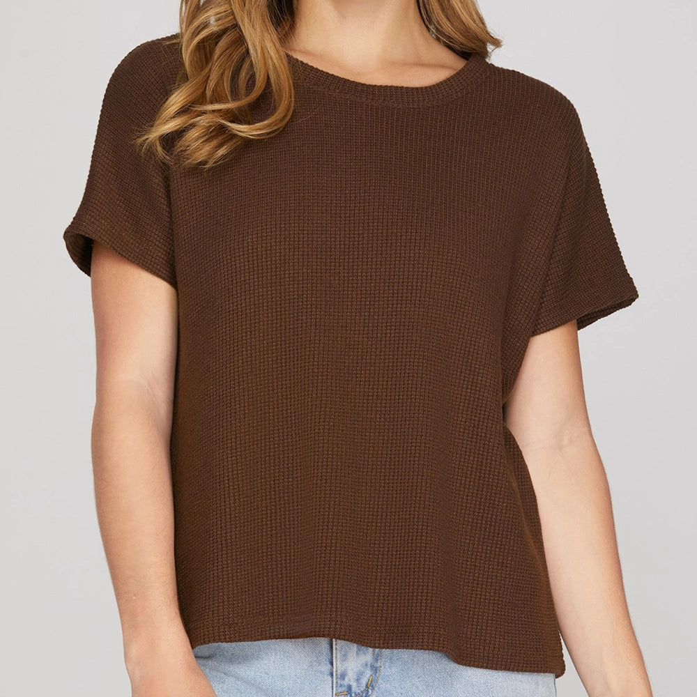 A woman wearing a dark brown, round-neck knit top with textured waffle fabric, drop sleeves, and blue jeans.