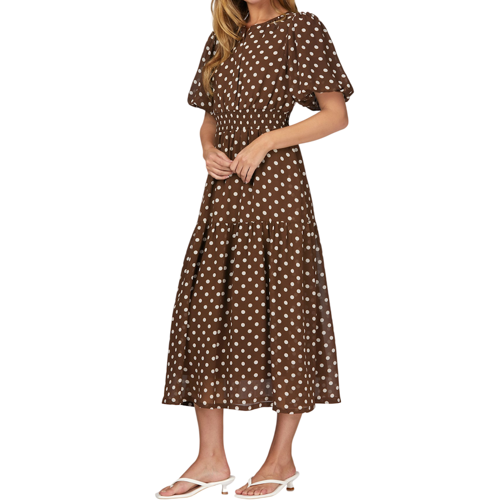 Woman in a brown polka dot midi dress with puff sleeves and a smocked waist, wearing white heeled sandals.