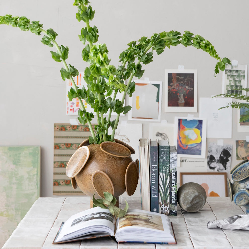 Green stalks in a textured brown footed vase with disc details, next to books on a tiled tabletop.