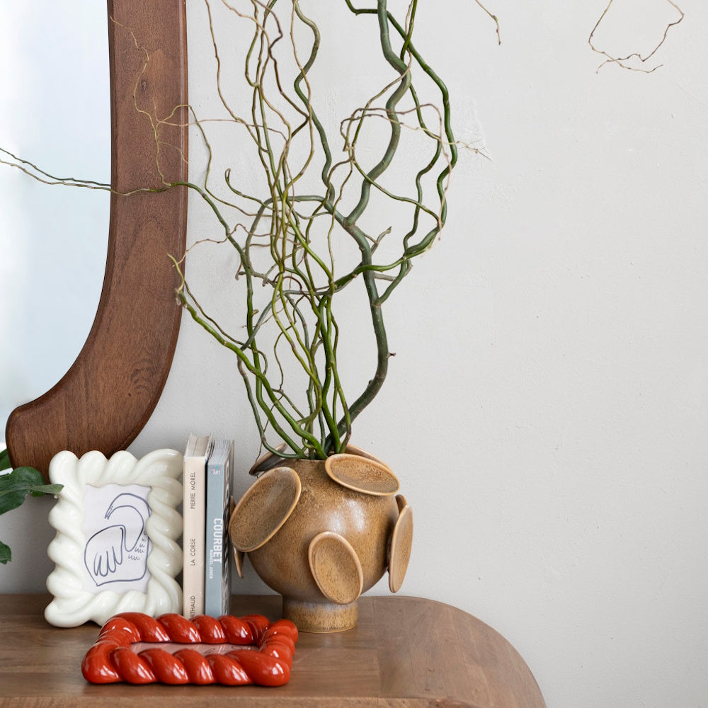 Brown spherical footed vase with 3D oval disks holding curly branches on a wooden table.