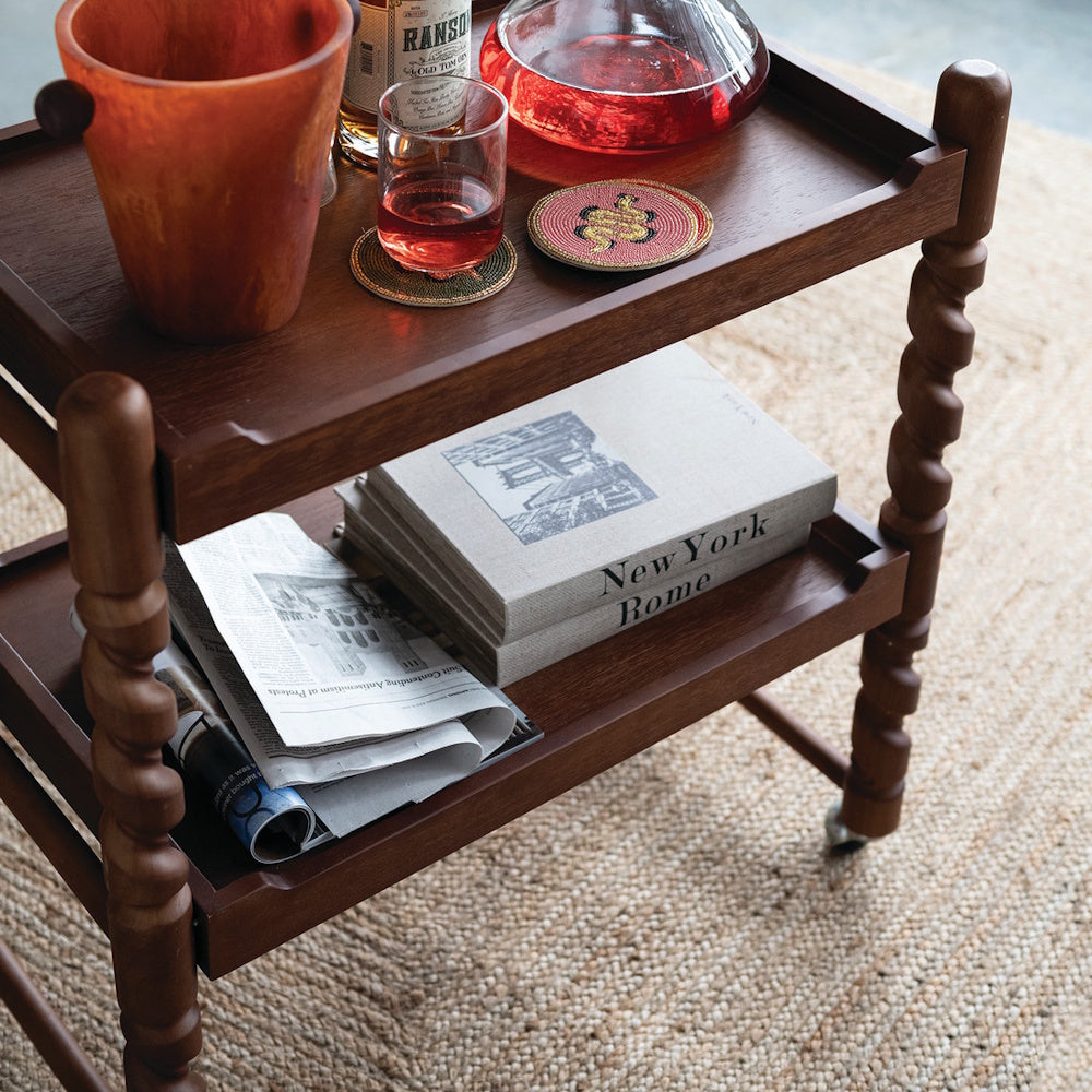 Wooden bar cart with spiral legs, decanter, cocktails, and coffee table books on a jute rug.