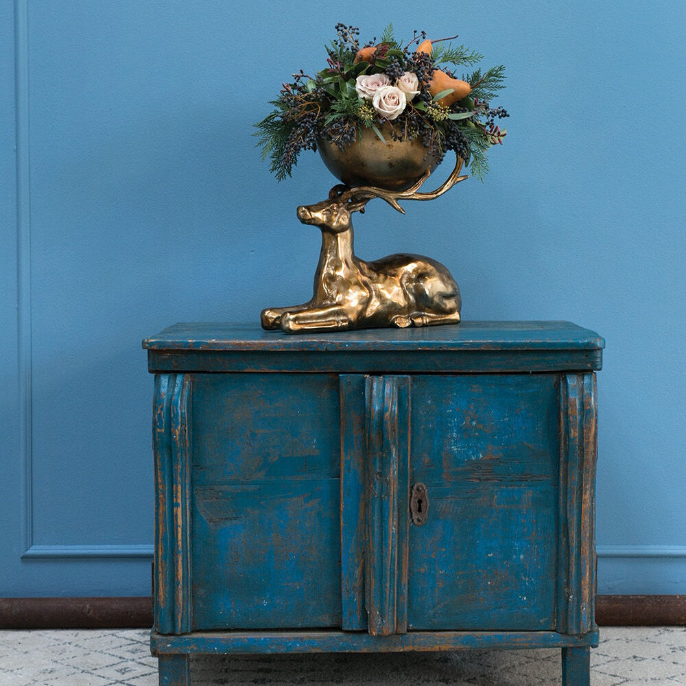 Brass deer-shaped bowl filled with a floral arrangement on a distressed blue wooden cabinet against a blue wall.