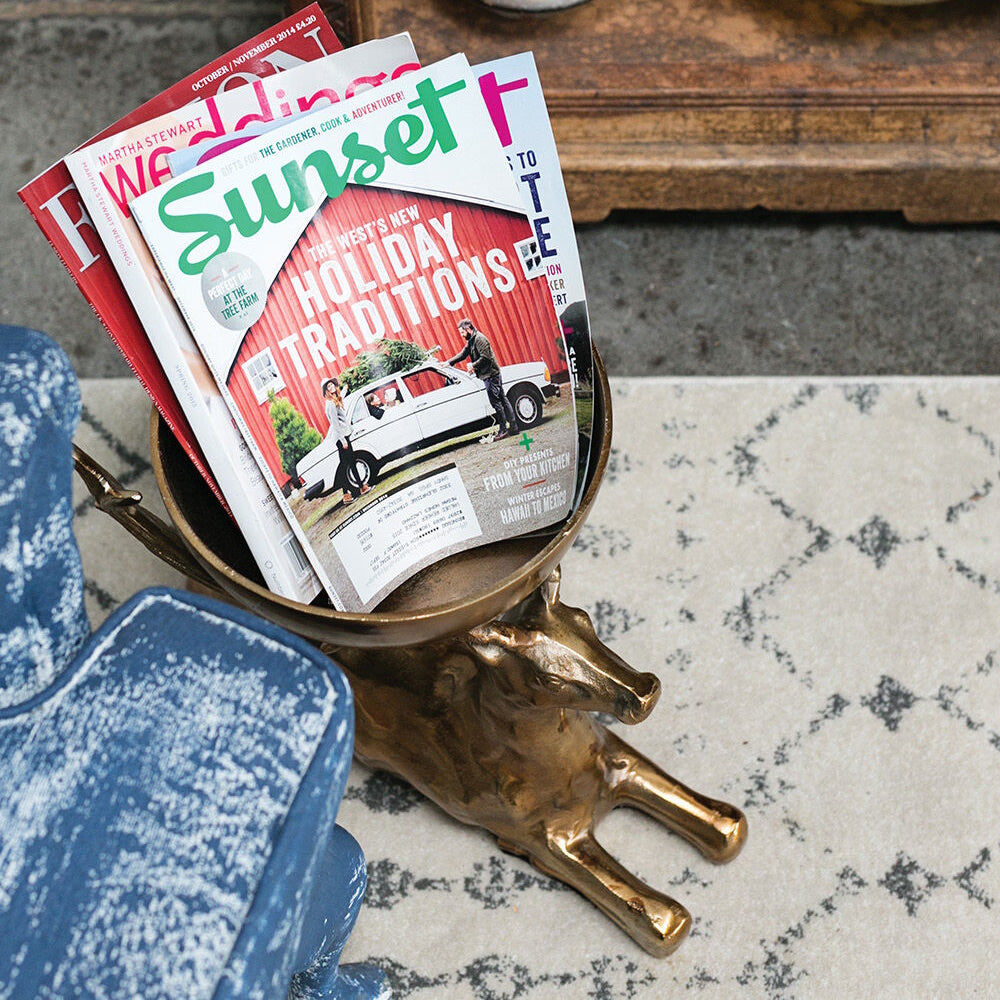 Brass deer-shaped pedestal bowl holding stacked magazines next to a blue patterned chair on a rug.