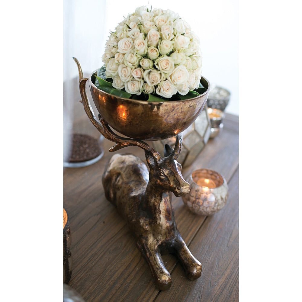 Bronze deer figurine bowl stand holding a dense arrangement of white roses on a wooden table.