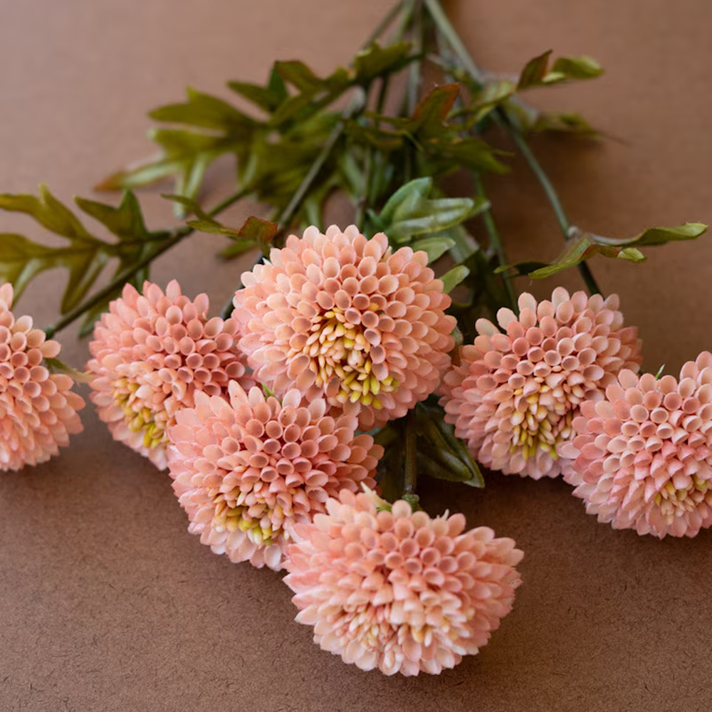 Bundle of seven artificial light pink ball dahlia flowers with green leaves on a brown textured surface.