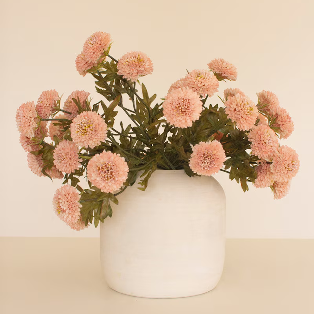 A bundle of light pink artificial ball dahlias in a round white ceramic vase on a neutral background.
