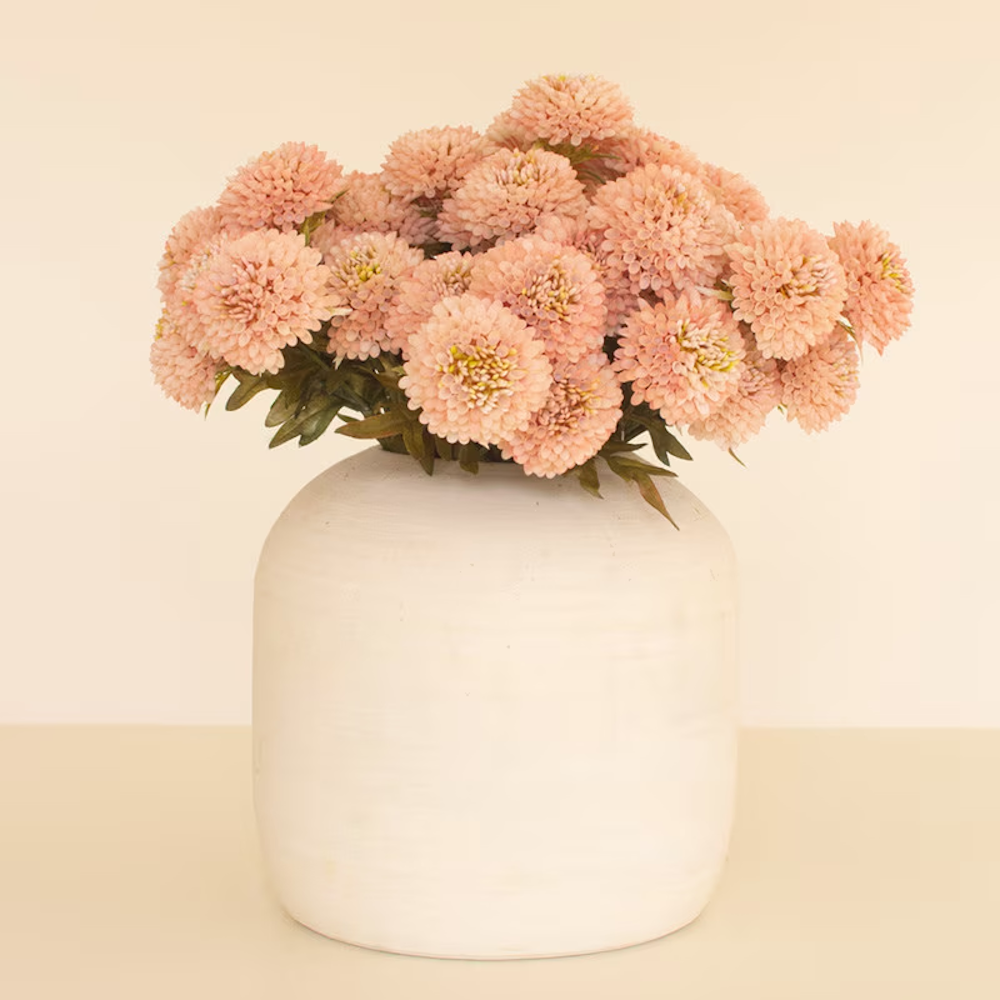 Bundle of light pink artificial ball dahlia flowers in a round white ceramic vase against a beige background.