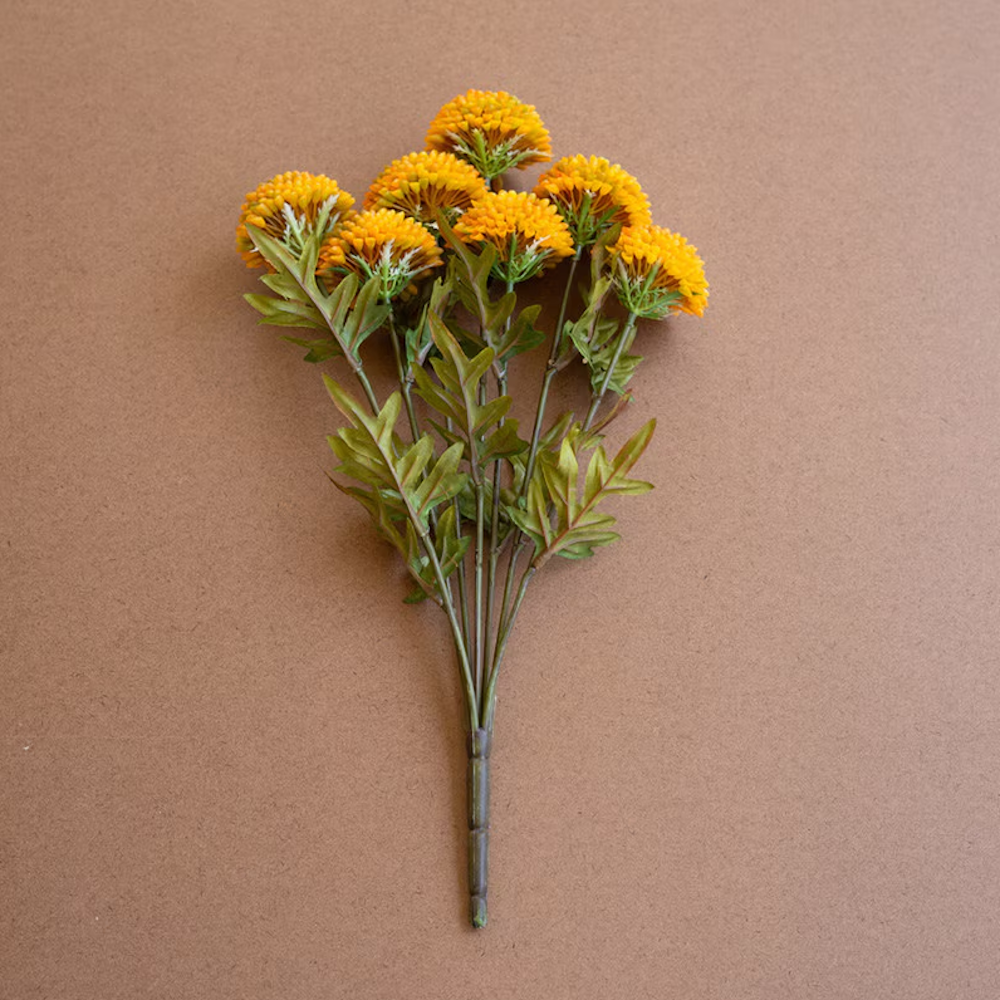 Bundle of artificial mustard yellow ball dahlias with green leaves lying flat on a brown backdrop.