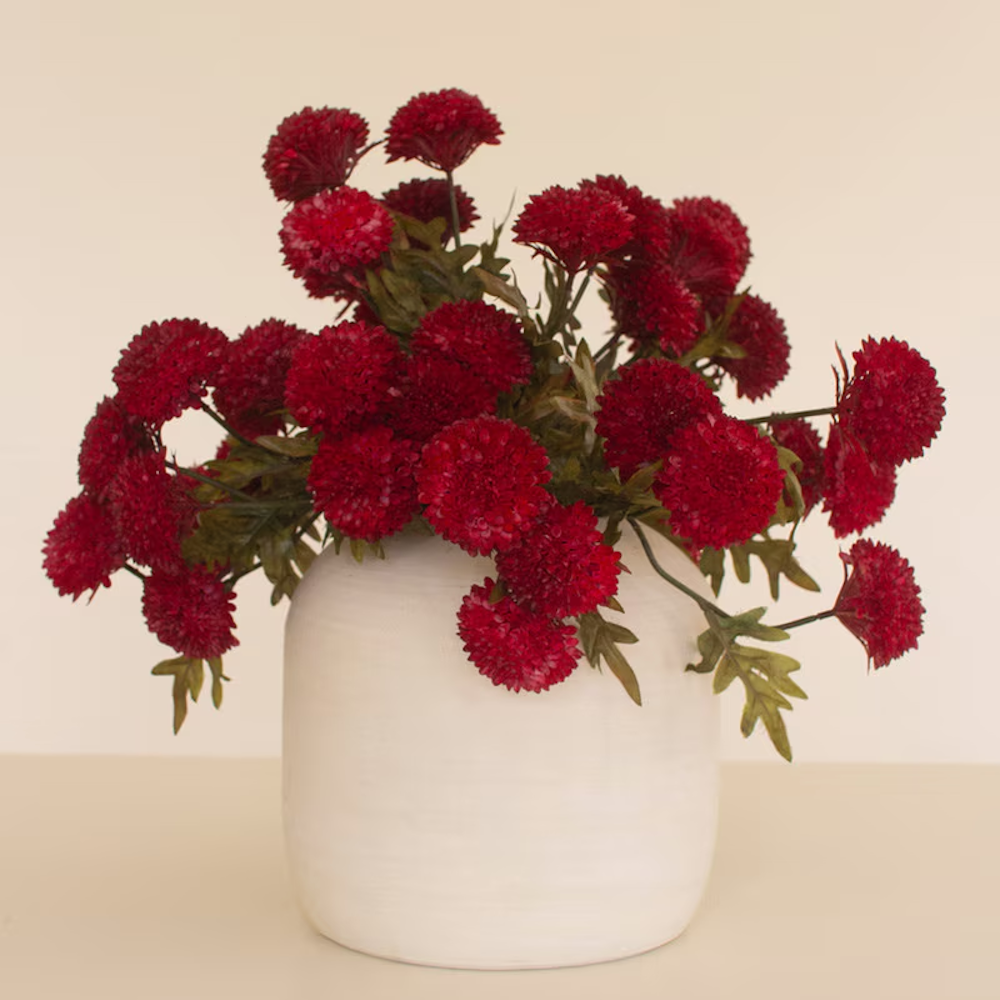 Full arrangement of deep red artificial ball dahlias in a white ceramic vase on a light backdrop.