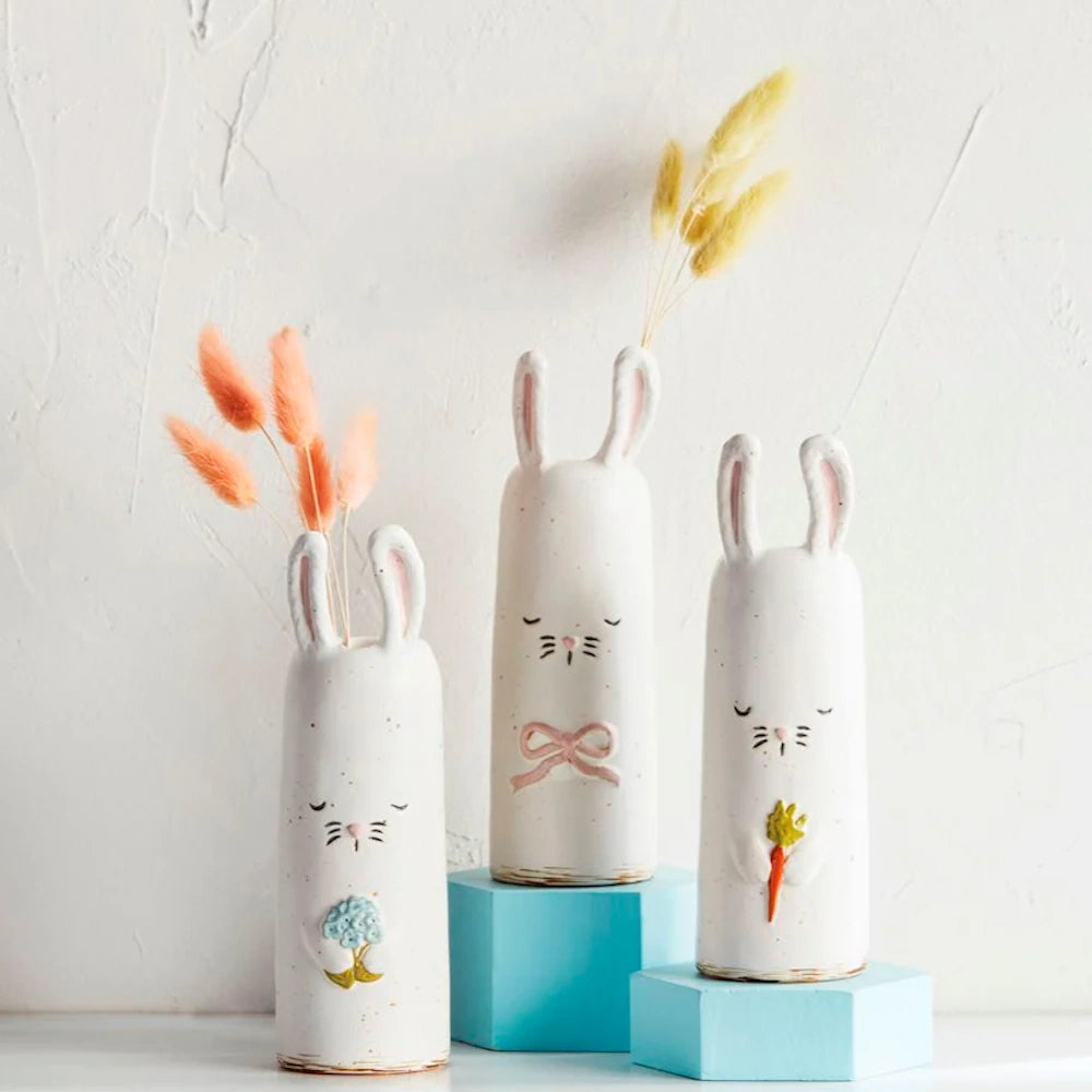 Three tall, white ceramic rabbit vases holding dried grasses on light blue display blocks.
