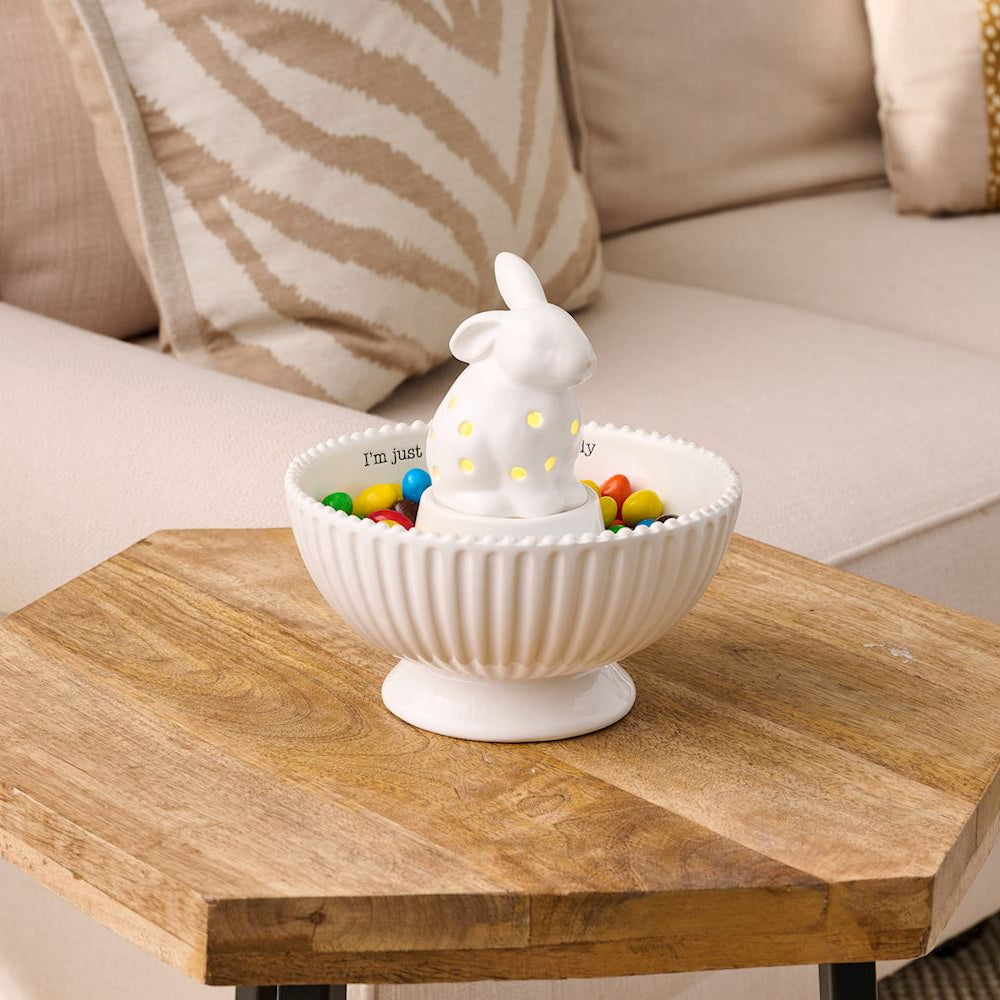 White ceramic light-up bunny sitter inside a fluted candy bowl filled with colorful chocolates on a wooden table.